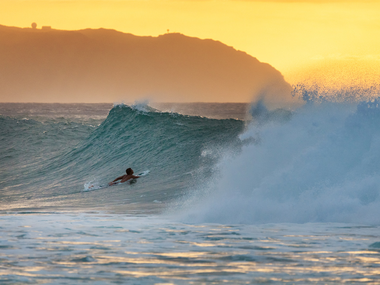 At sunset, a surfer paddes into a wave at banzai pipeline on Oahu's north shore.