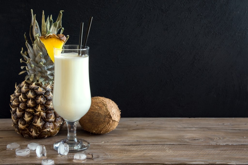 A piña colada, pineapple, and coconut sitting on a wooden table against a black background