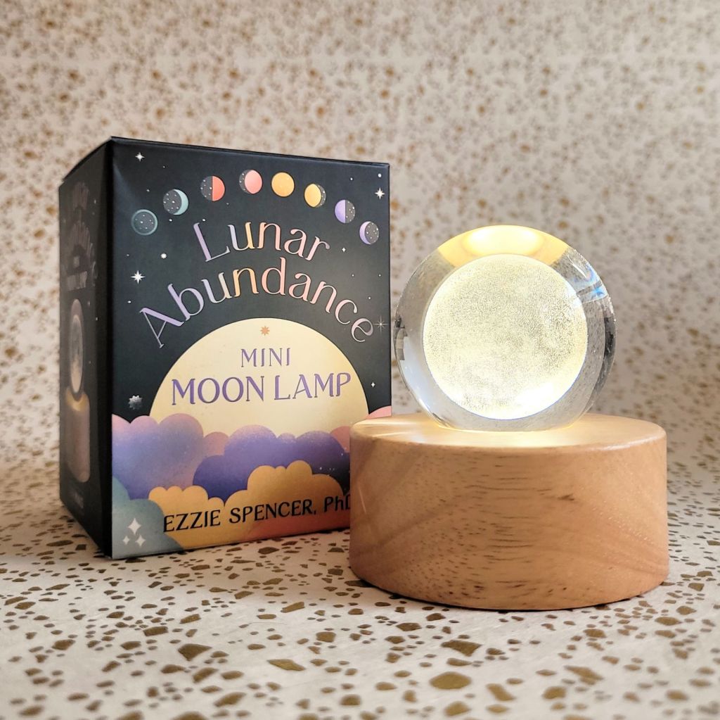 Photo of the “Lunar Abundance Mini Moon Lamp” box and included lamp and base laid above a gold and white backdrop. The lamp is lit.