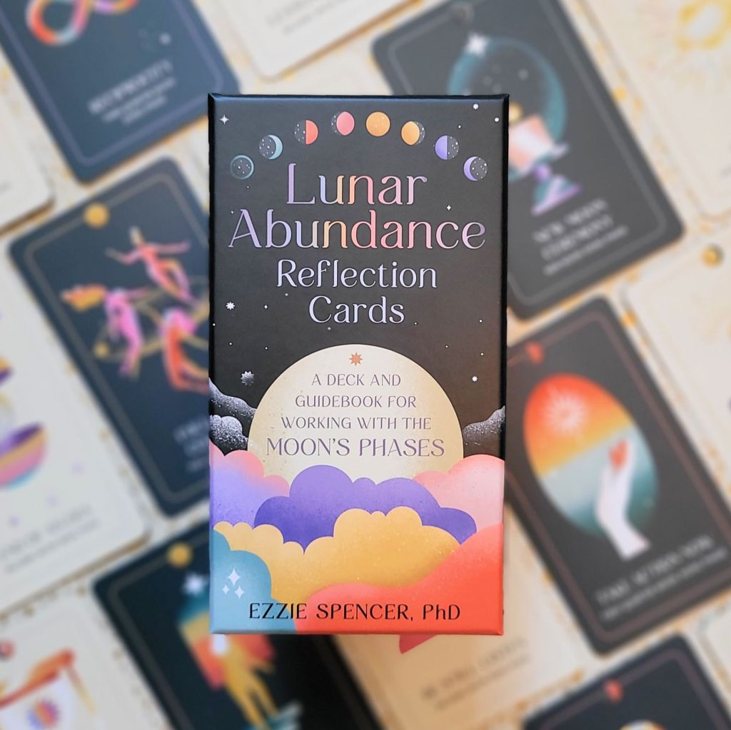 Photo of the “Lunar Abundance Reflection Cards: A Deck and Guidebook for Working with the Moon’s Phases” box laid above face-up cards from the deck, against a gold and white backdrop
