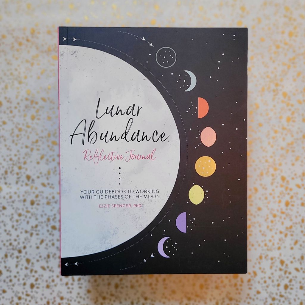 Photo of “Lunar Abundance: Reflective Journal: Your Guidebook to Working with the Phases of the Moon” laid above a gold and white backdrop