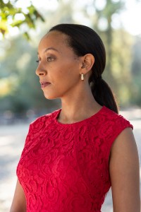 Haben Girma stands wearing a red dress with sunlit trees in the background.