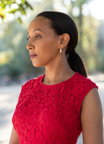 Haben Girma stands wearing a red dress with sunlit trees in the background.