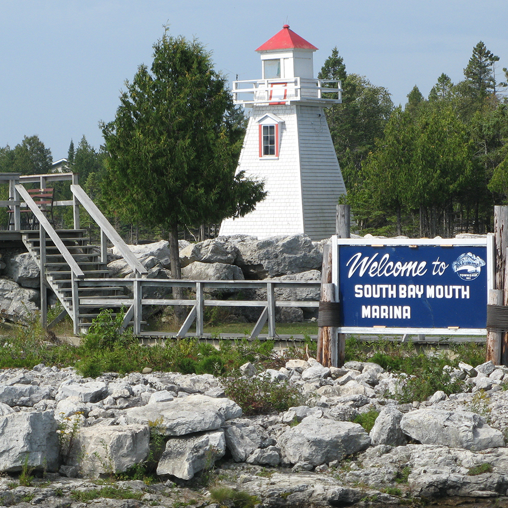 A lighthouse stands on a rocky and grassy island with a sign in front that reads Welcome to South Bay Mouth Marina