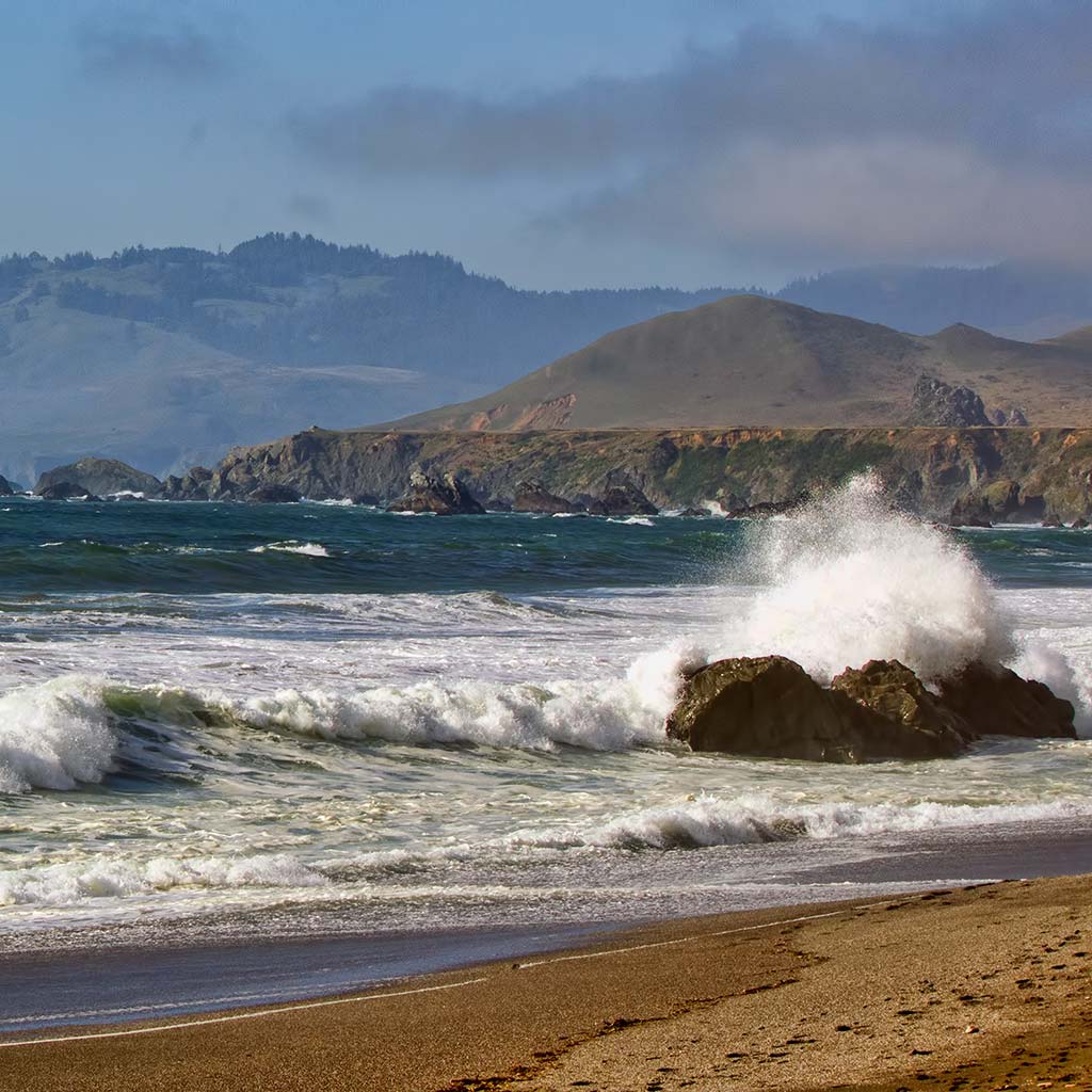 Looking out Wrights Beach towards the hills and cliffs in the distance. Waves crash onto shore and onto a boulder close to the shoreline