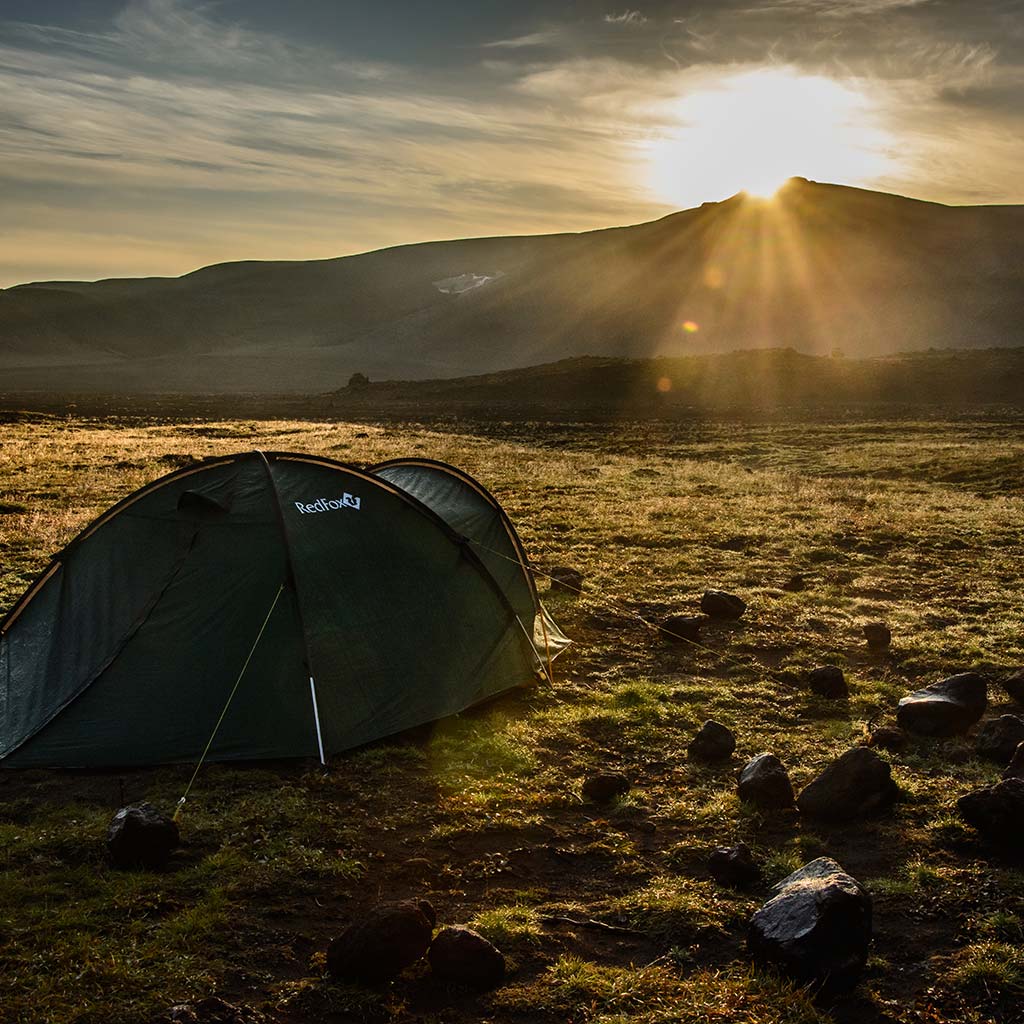 A tent in a field with the sun rising over a range of mountains.