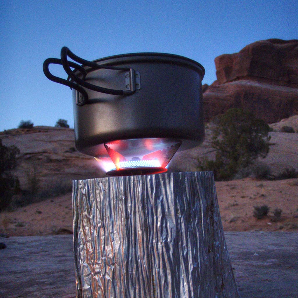 A portable stove lights a black cooking pot, the setup sits in front of the Canyonlands terrain at dusk