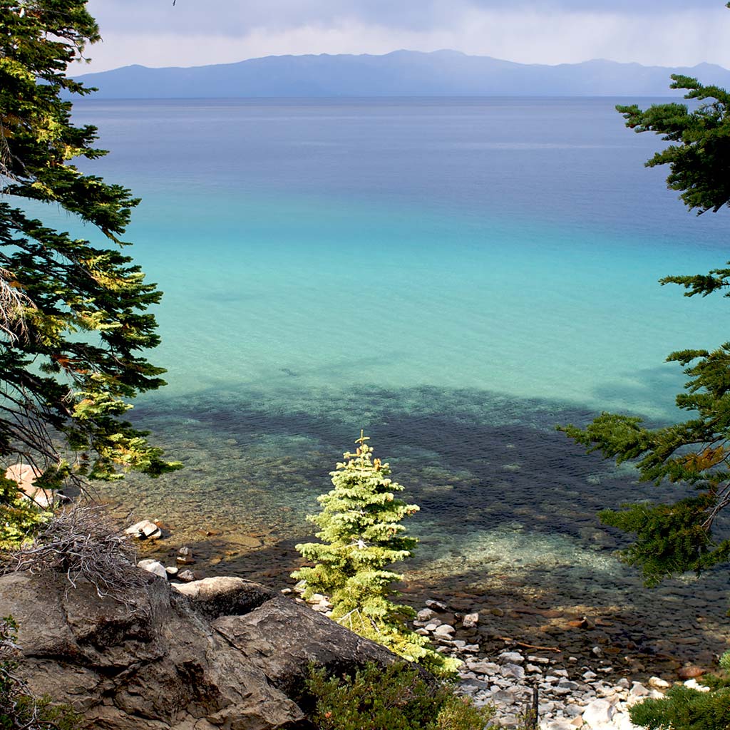 a boulder shoreline of dark rock and a few trees overlooks the bright blue water in DL Bliss State Park, with blue mountains looming across the water in the distance