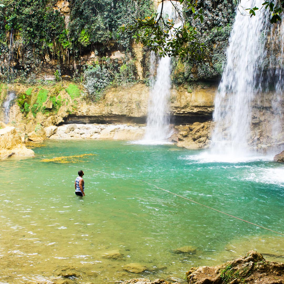 a man standing in green waters near 3 roaring waterfalls