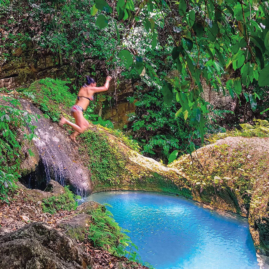 a woman jumping into a bright blue pool in a forest setting