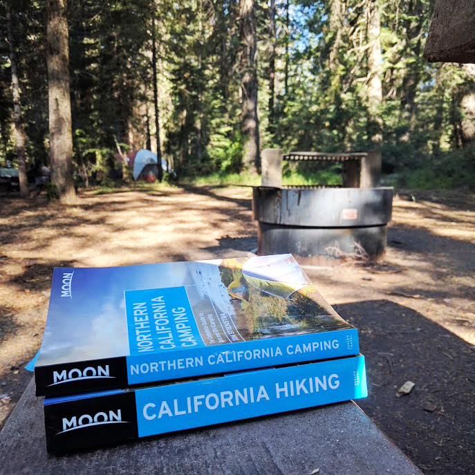Two Moon guidebooks (California Hiking and Northern California Camping) sit on a picnic table at Fowlers Camp, a fire pit and a distant tent are visible in the background