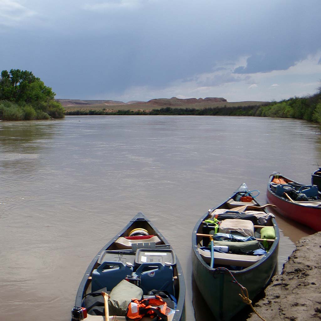 Canoes are parked and loaded with camping supplies on the bank of the Green River, looking out into the wide river and some hills in the distance