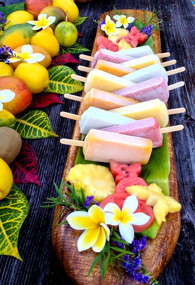 A wooden platter displays a colorful variety of frozen fruit pops, garnished with flowers and fruit.