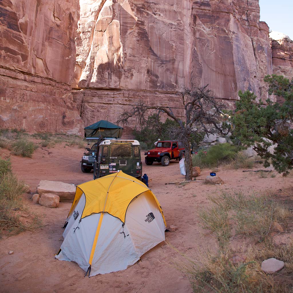 At the base of canyon rock formations sits a tent with a vehicle in the background 