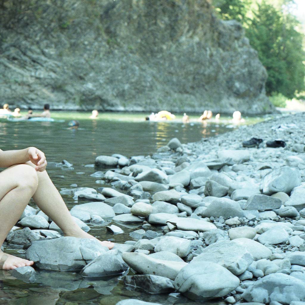 Sitting feet are pictured on the rocky shore of the Eel River, in the background people are swimming in the river.