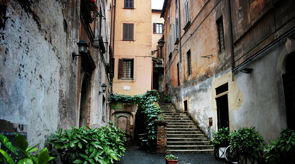 Plants and ivy line a narrow street in Rome with stone steps leading up to a second level between buildings.