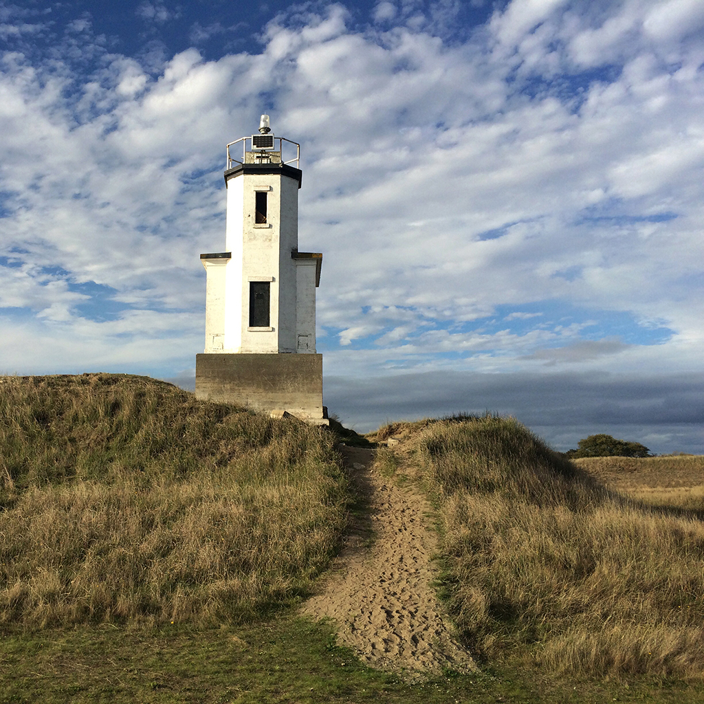 A lighthouse stands on a grassy hill with a sandy path leading up to it and a blue sky with clouds in the background