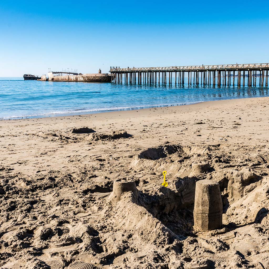 A sandcastle on the beach on a clear day with a pier and the shipwreck in the background