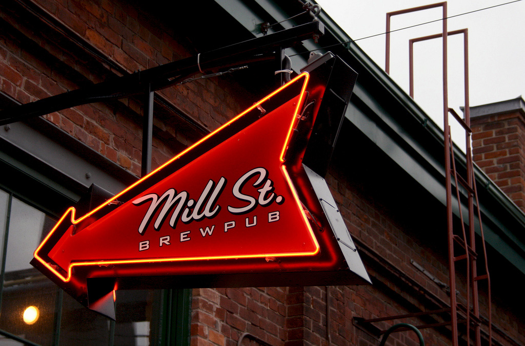 Outside a brick warehouse building, an arrow shaped neon sign will Mill St Brewery logo.