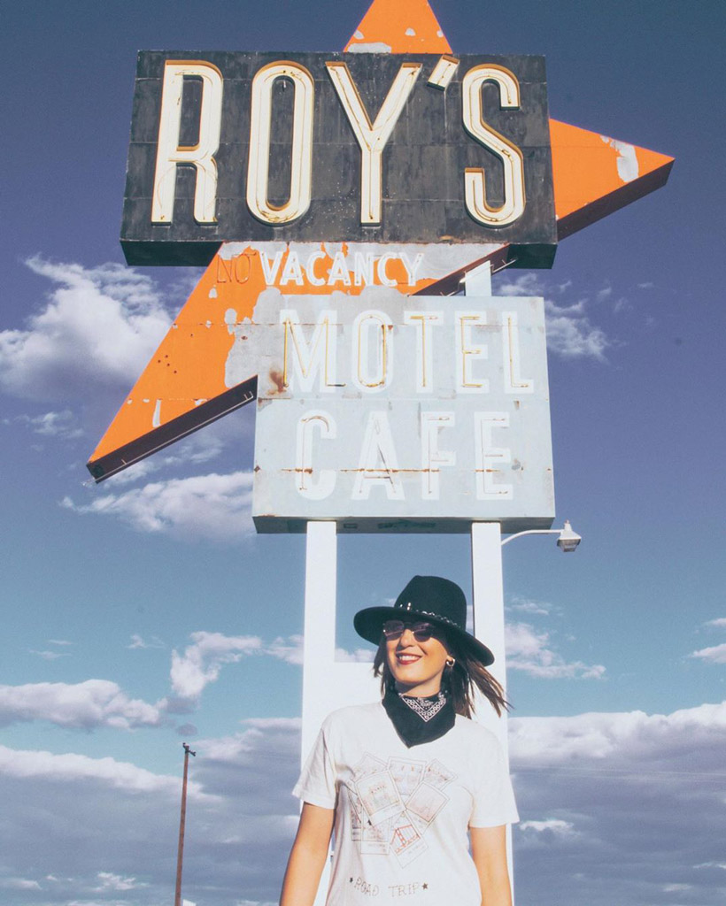 woman in a black hat posing in front of a faded hotel sign on route 66 with blue sky in the background