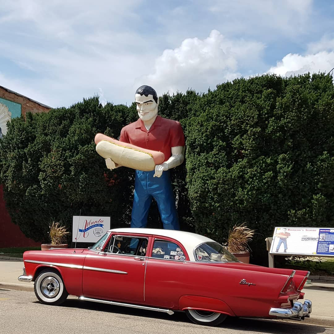 statue of a man holding a hotdog with a red classic car parked in front