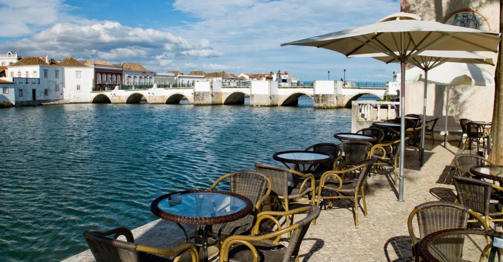 Roman bridge in the background, with cafe tables and umbrellas beside the Gilao river in Tavira, Algarve, Portugal.