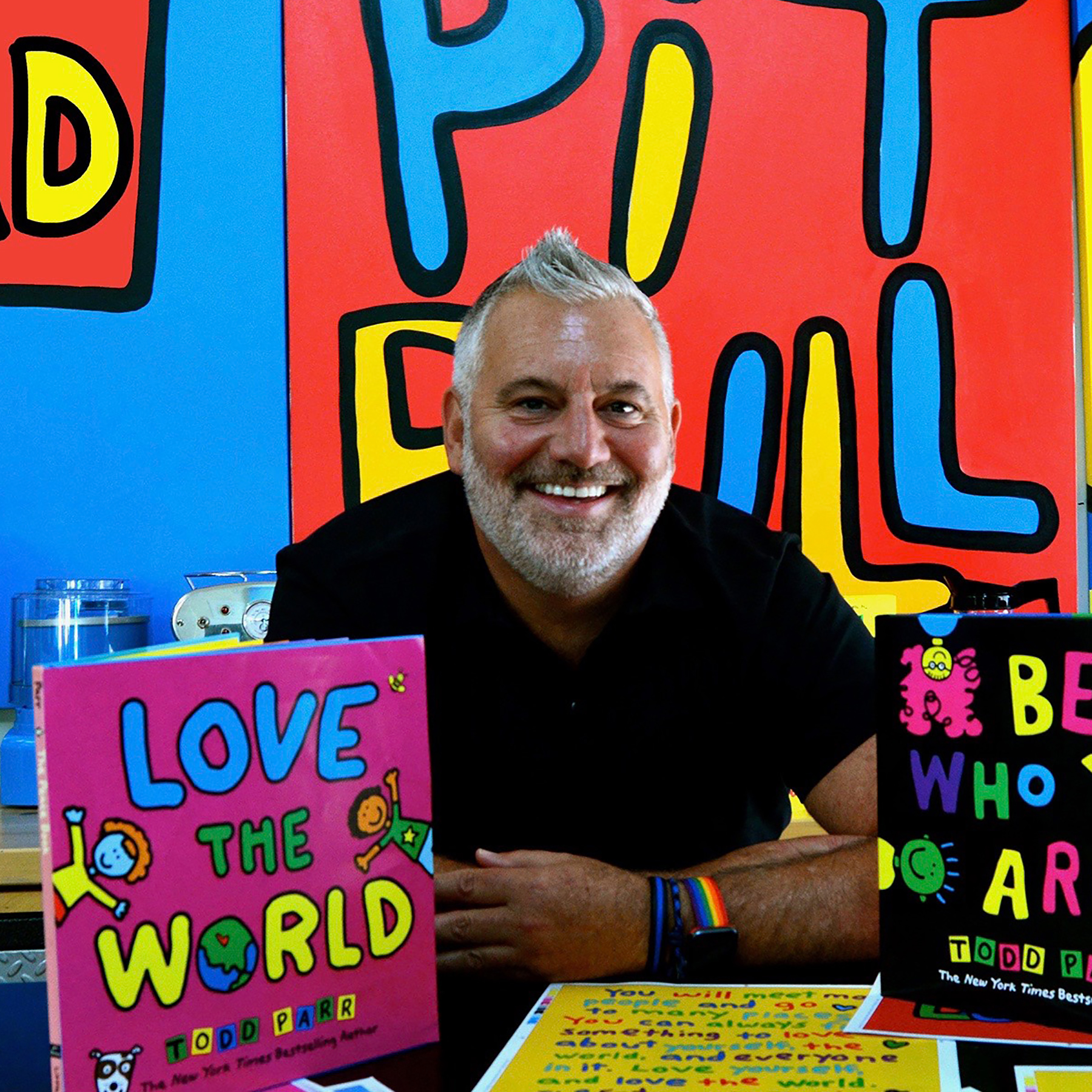 Image of Todd Parr sitting at a table with his books "Love the World" and "Be Who You Are"
