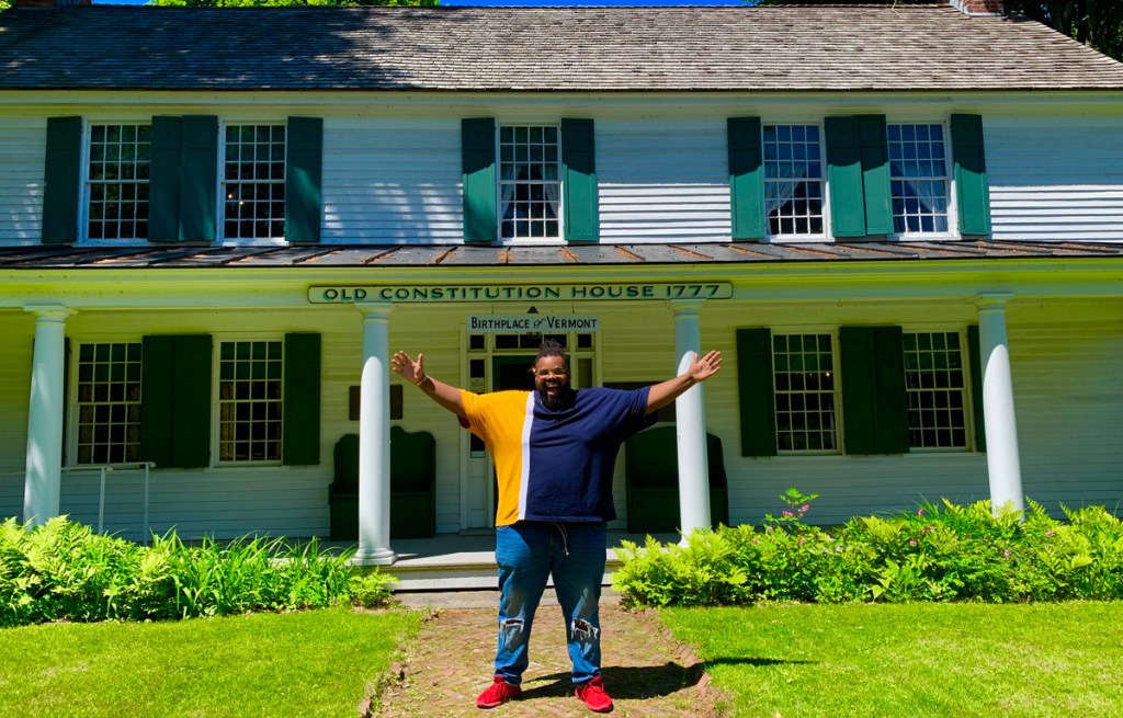 a Black plus-size man spreading his arms wide standing in from of an historic building in windsor, vermont
