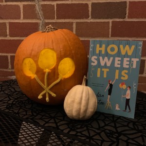 Pumpkin with balloons carved in it near a copy of How Sweet It Is by Dylan Newton, near a tiny white pumpkin.