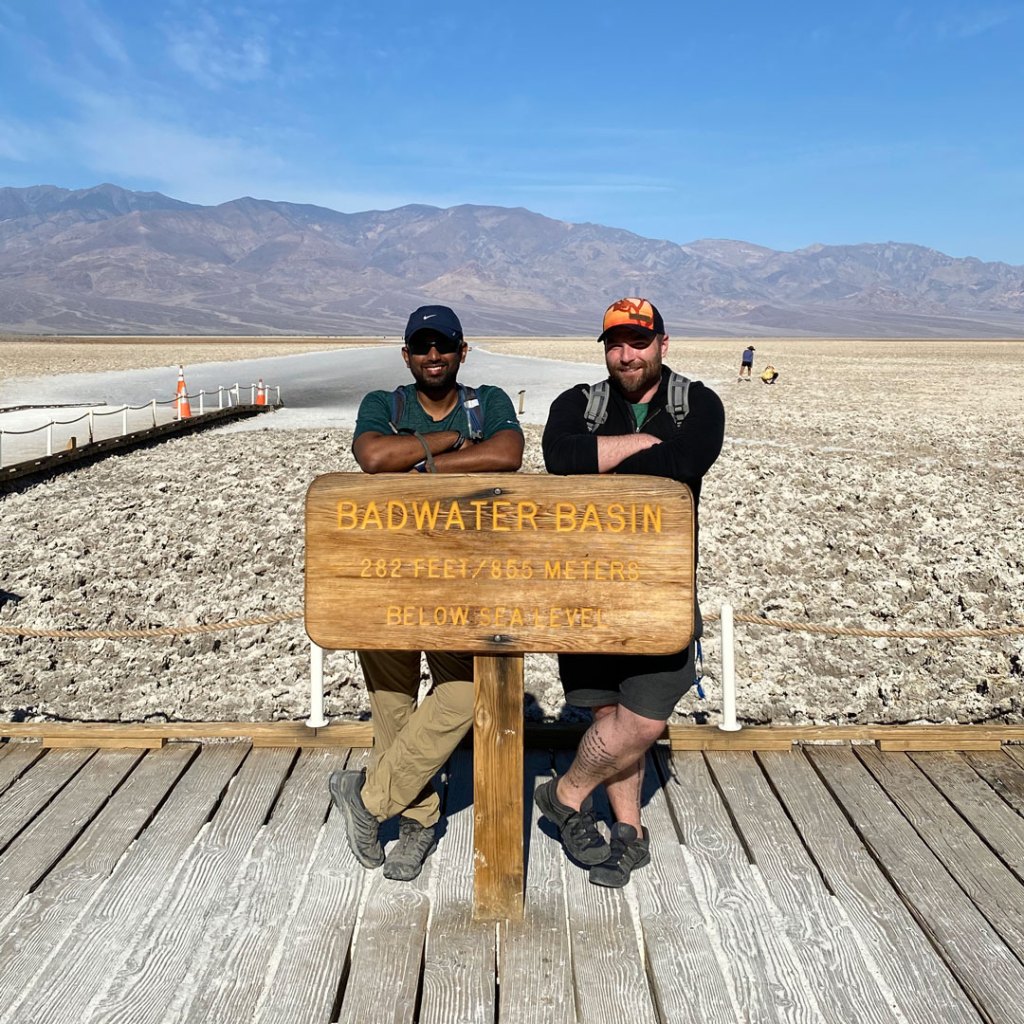 two men, one dark-complected and one light-complected, wearing hats and smiling while posing behind a wooden sign that says Badwater Basin with desert in the background