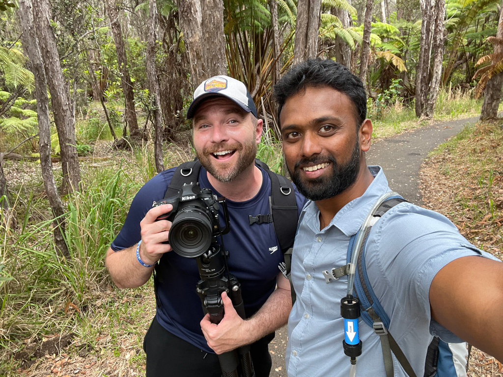 selfie of one bearded light-complected man smiling while holding a camera next to a bearded, medium-brown-complected man smiling with trees in the background