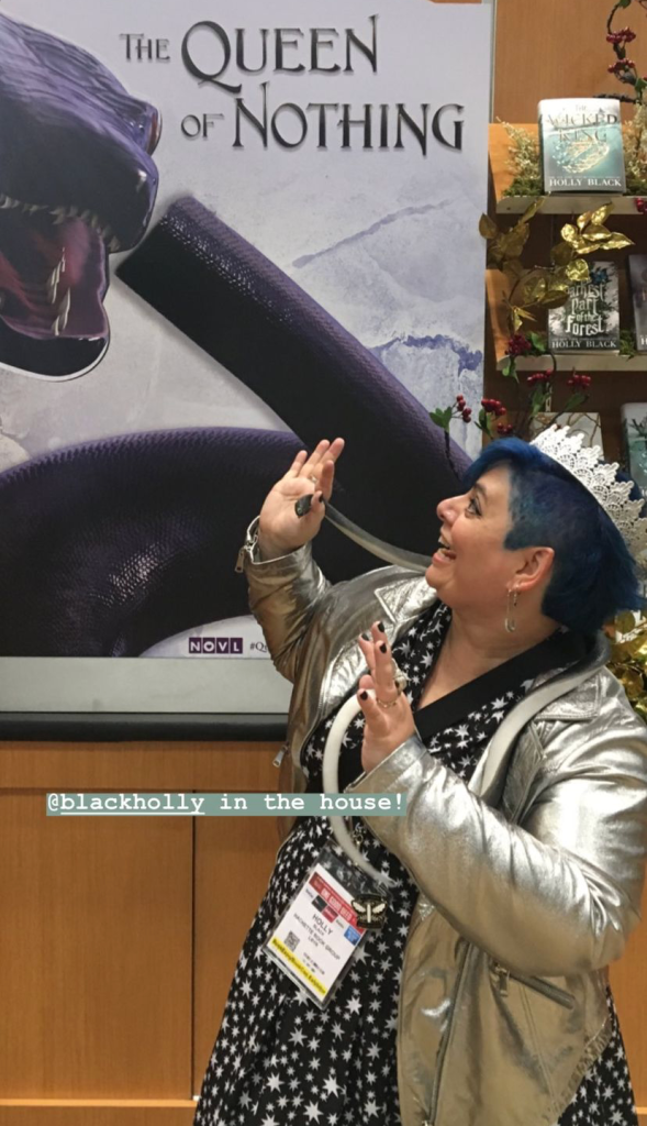 NOVL - Image of Holly Black standing in front ofa poster of her book 'The Queen of Nothing', pretending to dodge a snake on the cover