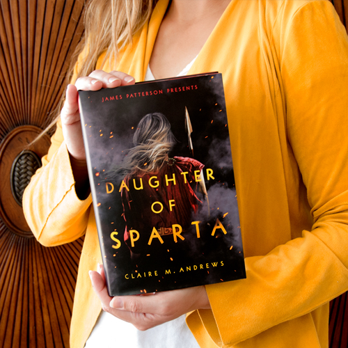 NOVL - Instagram image of person in yellow cardigan holding up book cover for 'Daughter of Sparta' by Claire M. Andrews