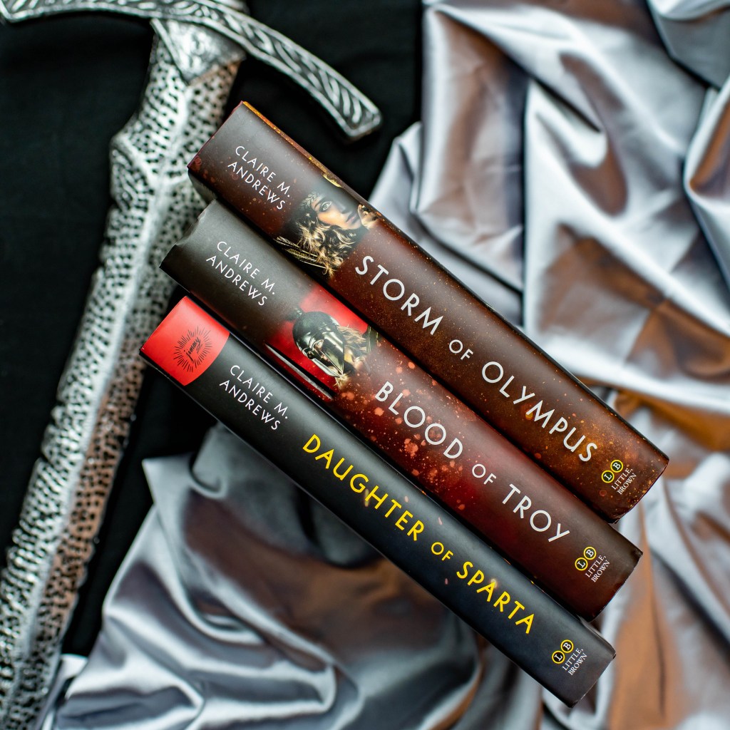 Image of the hardcover spines of the books "Daughter of Sparta", "Blood of Troy", and "Storm of Olympus" by Claire M. Andrews