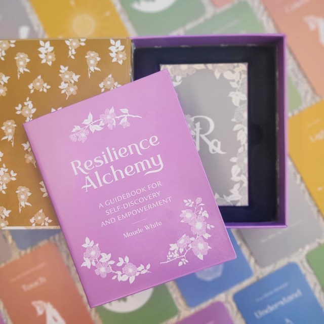 Photo of an open box of "Resilience Alchemy" showing the guidebook laid next to the box of cards, all laid above face-up cards from the deck