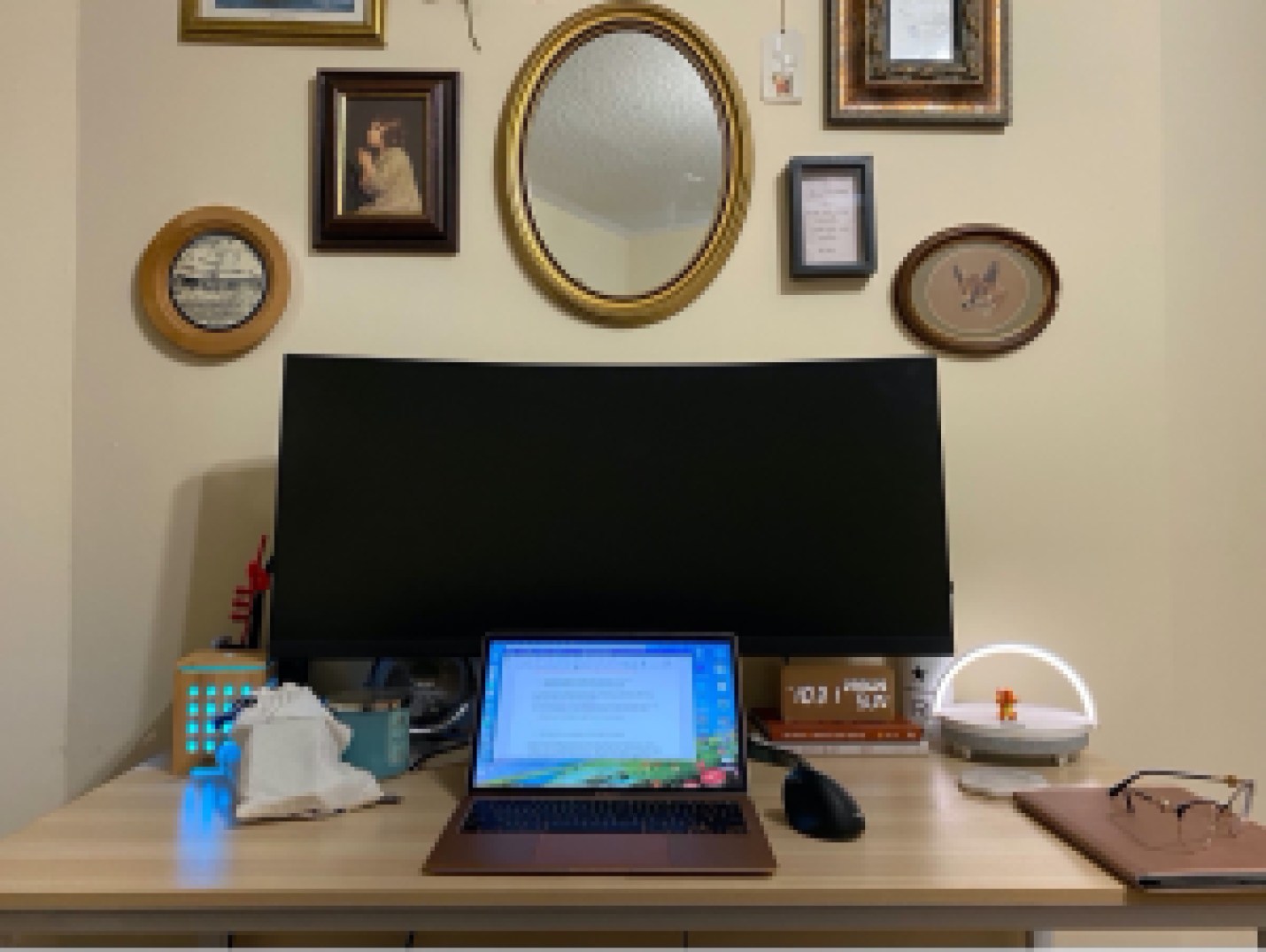 A view of a desk with a laptop in the foreground and a monitor behind. On the wall there are framed photos and a mirror. 