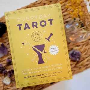 Photo of the revised paperback edition of "Everyday Tarot" laid on a woven mat next to flowers and crystals