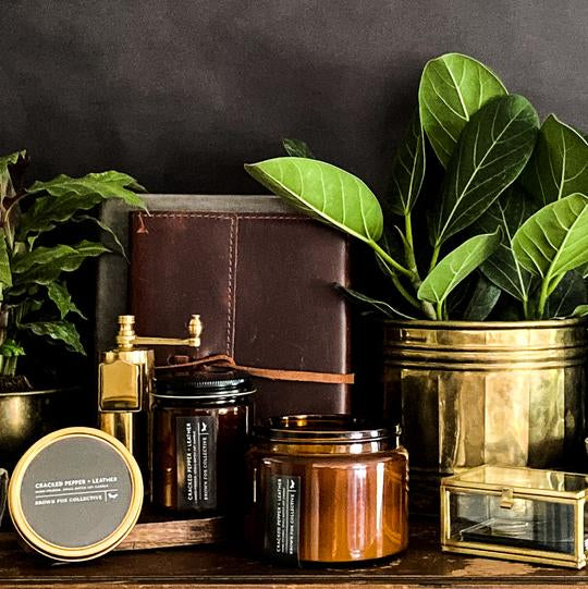 Two brown glass candle jars sit on a wooden shelf among other objects including a brown leather diary, glass box, and two green houseplants