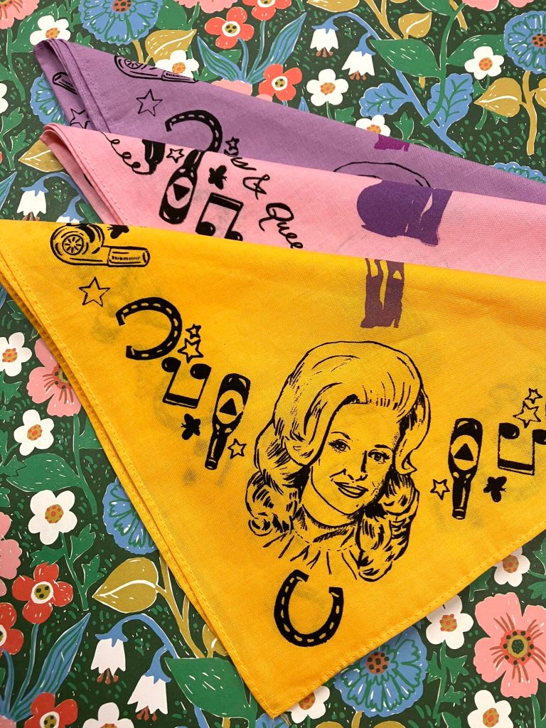 Three folded overlapping yellow, pink, and purple bandanas feature a printed line drawing of Dolly Parton against the background of a colorful flowerful print