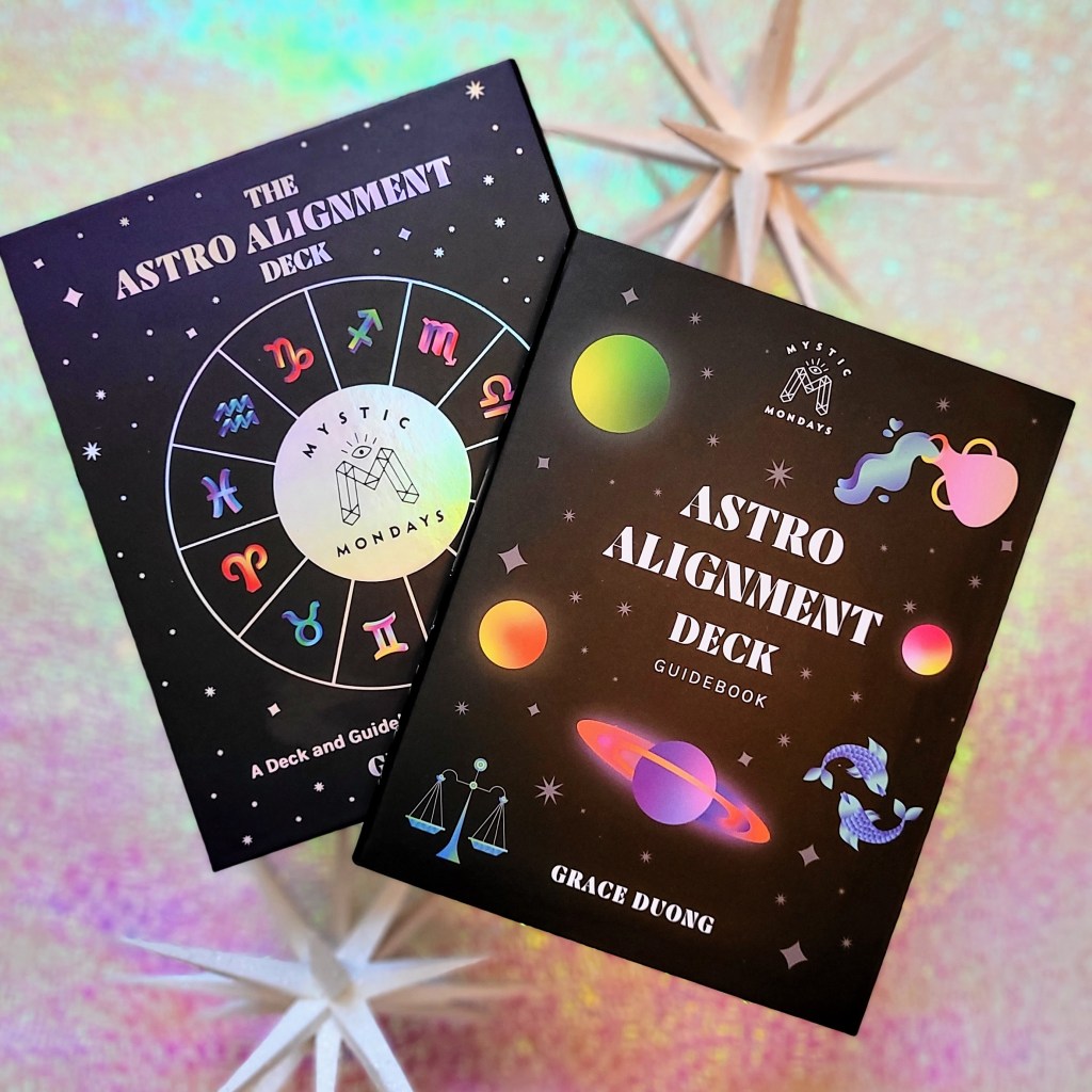 Photo of "Mystic Mondays: The Astro Alignment Deck" box and guidebook laid next to decorative stars above a pink and green iridescent background.