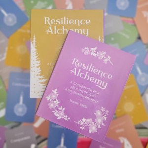 Photo of the box and guidebook of "Resilience Alchemy" laying above artfully scattered cards from the deck.