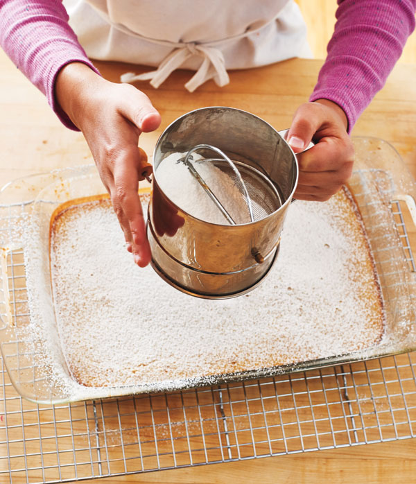 Photo of hands sprinkling powdered sugar over lemon squares.