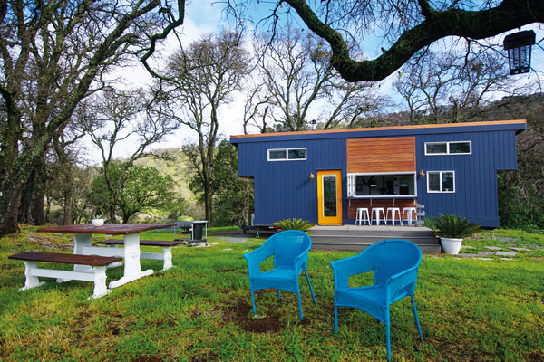 Photo of the exterior of a blue tiny house and front yard with a picnic table and two blue chairs.