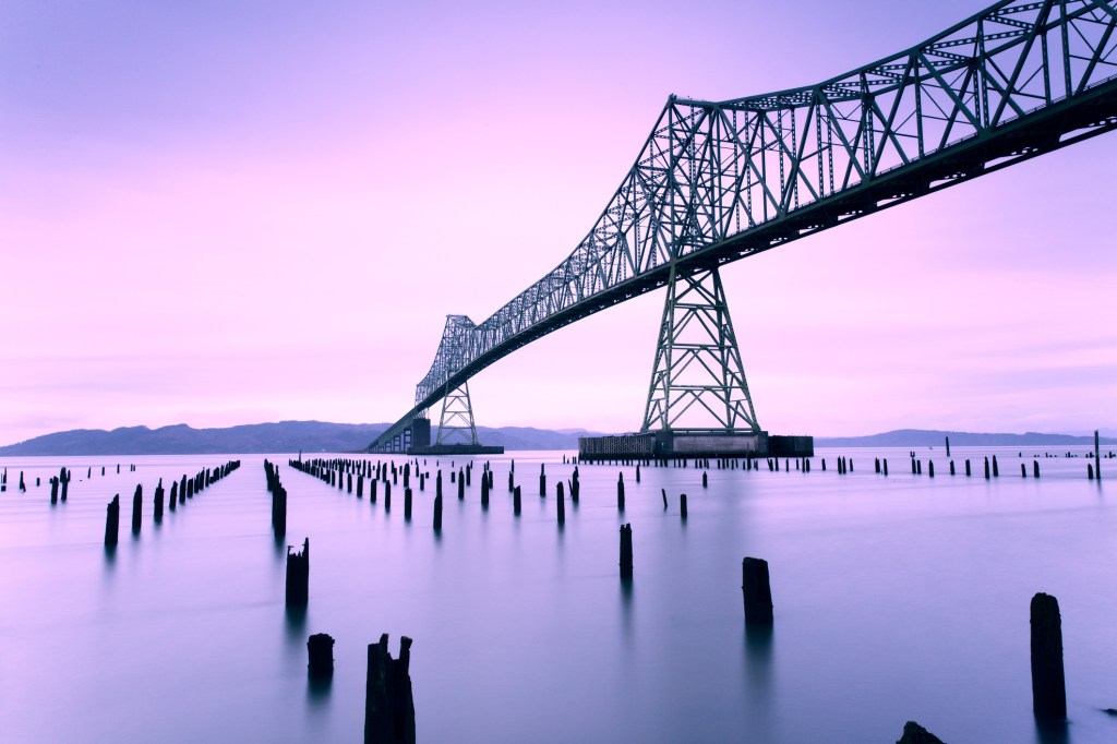 Image of steel bridge over river with purple sunset reflecting in the water.