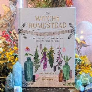 Photo of "The Witchy Homestead" standing among flowers and various green crystals