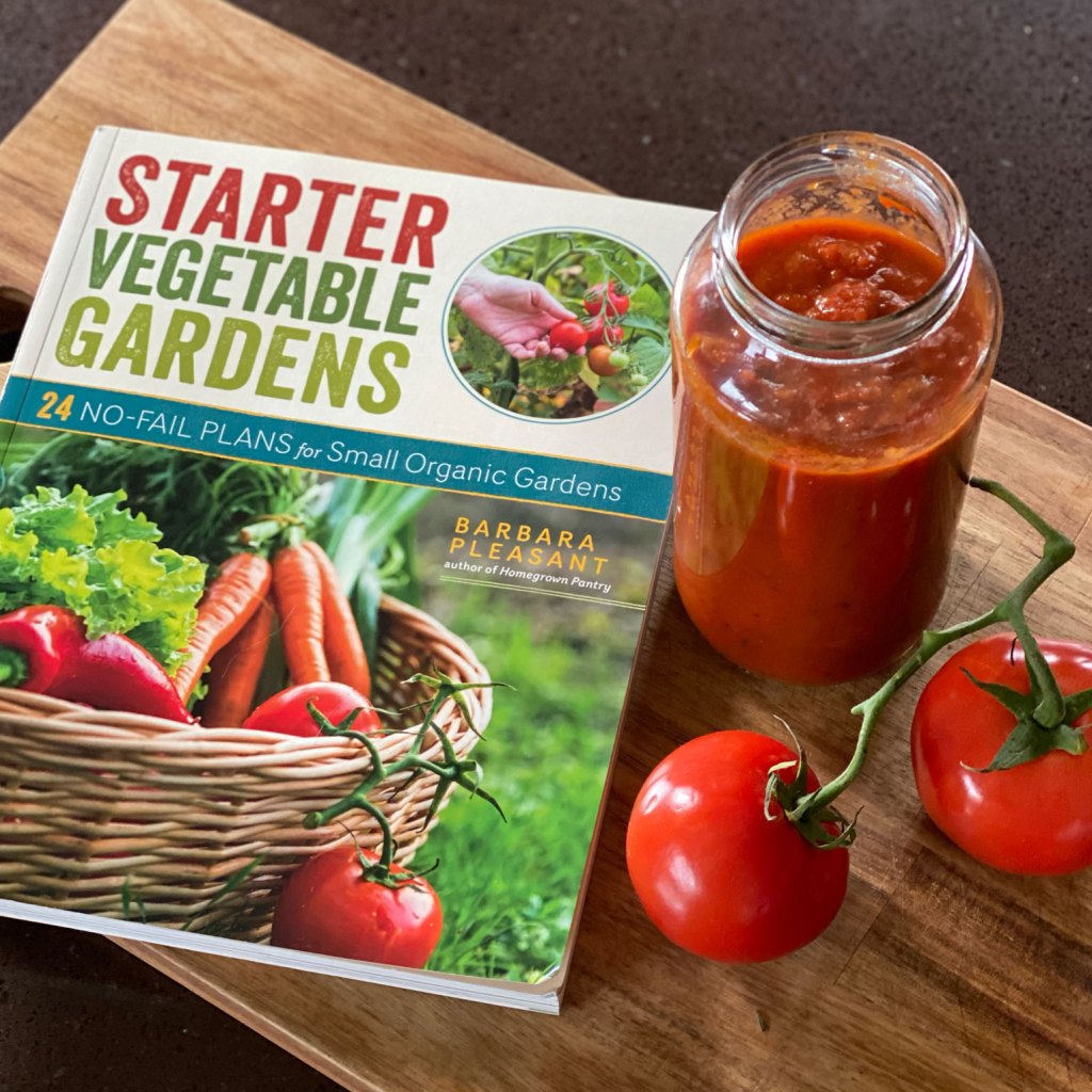 A photo of the Starter Vegetable Gardens book with a jar of sauce and tomatoes.