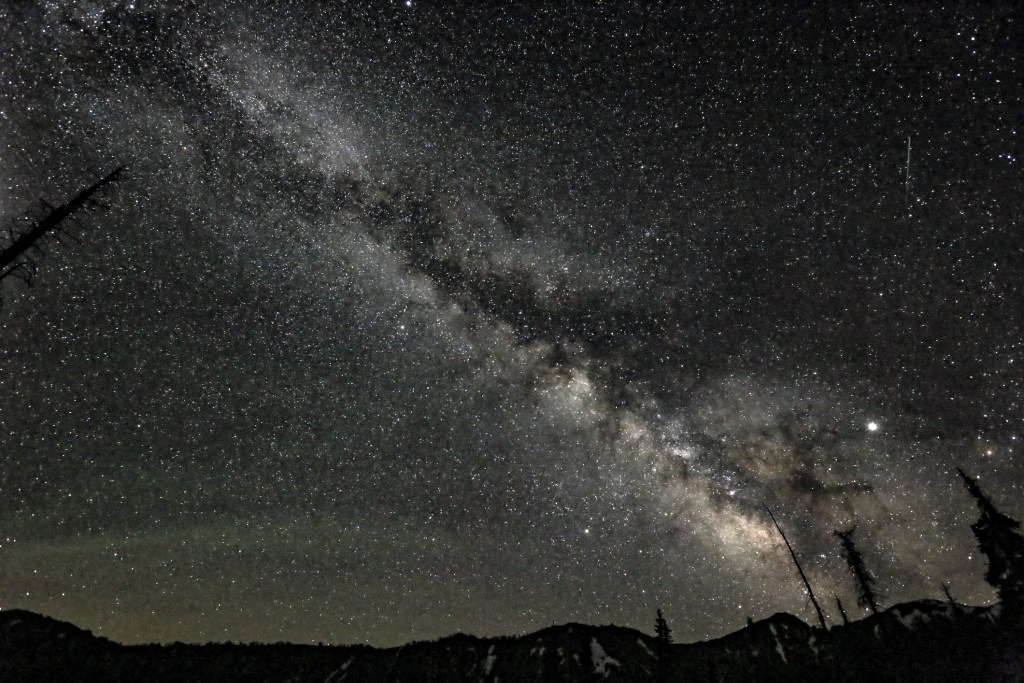 A galaxy stretches out across a night sky filled with white stars over the silhouettes of mountains.