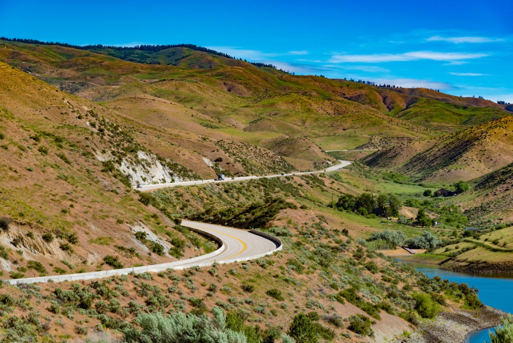 A winding road leads through rolling brown and green hills next to a river under bright blue sky.