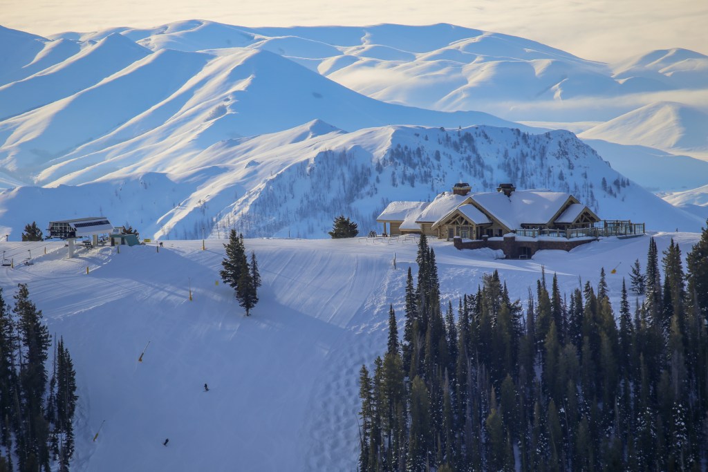 A ski chalet perched at the top of a ski hill with pine trees and rolling snow-covered mountains in the distance.