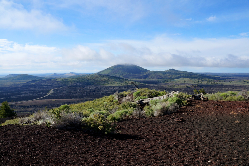 Black volcanic dirt in the foreground of a vista overlooking rolling green and black hills. 
